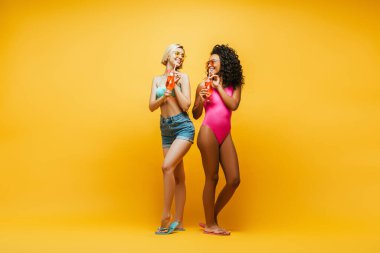 Sarıda kokteyl bardakları tutarken birbirine bakan ırklar arası genç arkadaşların tam boy görüntüsü.