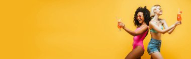 Sırt sırta duran, sarıya izole edilmiş kokteyl bardaklarıyla yaz kıyafetli genç çok kültürlü kadınların yatay görüntüsü.