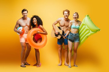 Gitarlı, şişme toplu, şilteli ve yüzme yüzüklü genç ve çok kültürlü arkadaşların sarı kameraya bakışları.