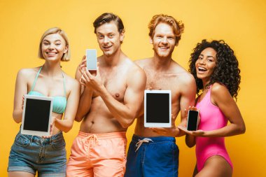 Yazlık kıyafetli genç çok kültürlü arkadaşlar sarı ekranlı cihazlar gösteriyorlar.