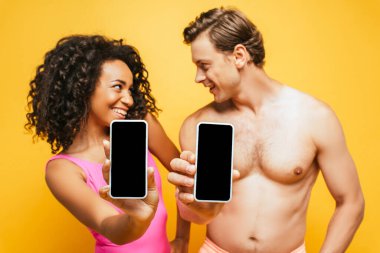 Kıvırcık Afrikalı Amerikalı kadın ve tişörtsüz adam birbirlerine bakarken sarı ekranda boş ekranlı akıllı telefonlar gösteriyorlar.