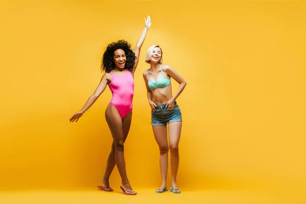 兴奋的非洲裔美国女人高举双手 金发碧眼的朋友手插在黄色口袋里的全景 — 图库照片