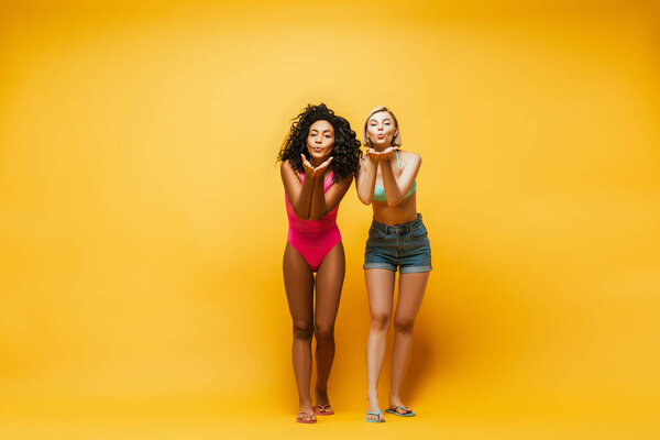 full length view of multicultural women blowing air kisses at camera on yellow