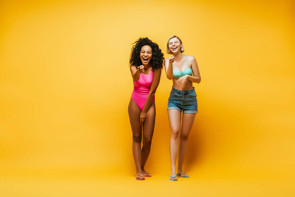 full length view of excited interracial women laughing and pointing with fingers at camera on yellow