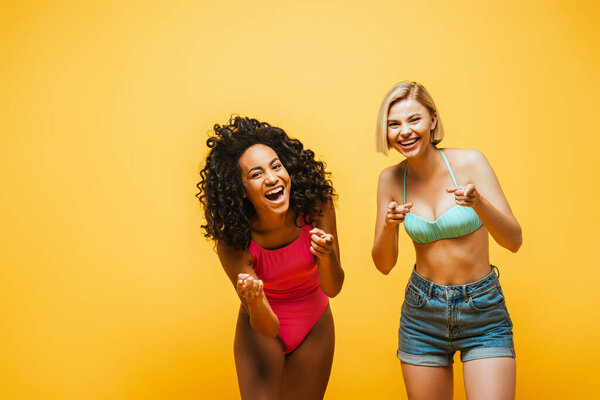 excited interracial women in summer outfit laughing and pointing ith fingers at camera isolated on yellow