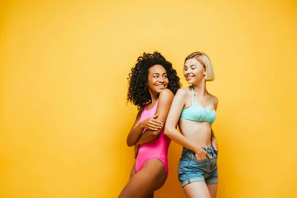 african american women with crossed arms and her blonde friend with hands in pockets standing back to back on yellow