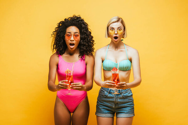 excited interracial women in summer outfit holding cocktail glasses and looking at camera on yellow