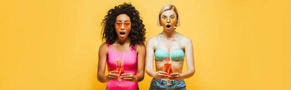 horizontal image of shocked interracial women in summer outfit holding cocktails and looking at camera isolated on yellow