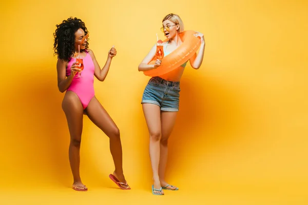 身穿泳衣的年轻的非洲裔美国女人 戴着泳圈的金发碧眼的朋友 戴着黄色的鸡尾酒杯 — 图库照片