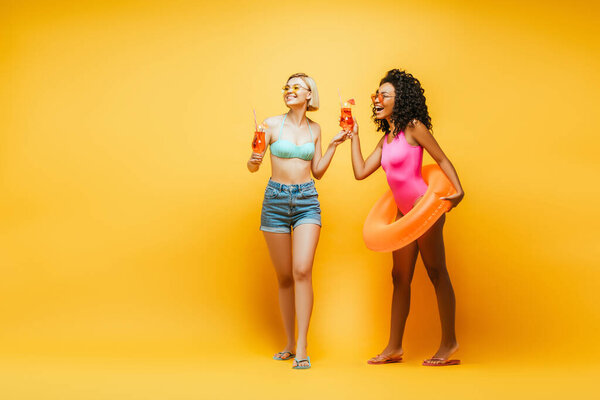 blonde woman with african american friend holding cocktail glasses and swim ring laughing while looking away on yellow