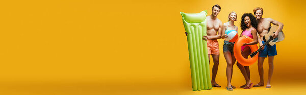 горизонтальное изображение мультикультурных друзей с гитарой, надувным матрасом, мячом и кольцом для плавания, смотрящих в камеру на желтый