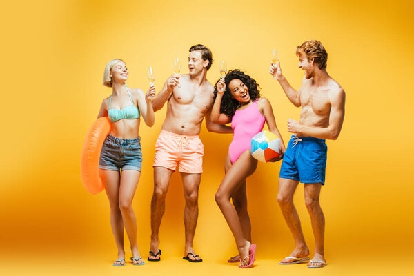 young multicultural friends in summer outfit holding champagne glasses, inflatable ball and ring on yellow