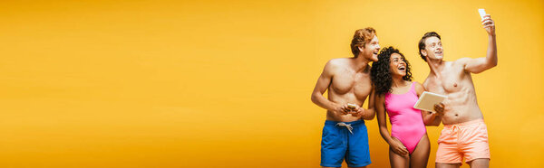 горизонтальное изображение молодого человека, делающего селфи на смартфоне с мультикультурными друзьями, держащими гаджеты изолированные на желтом