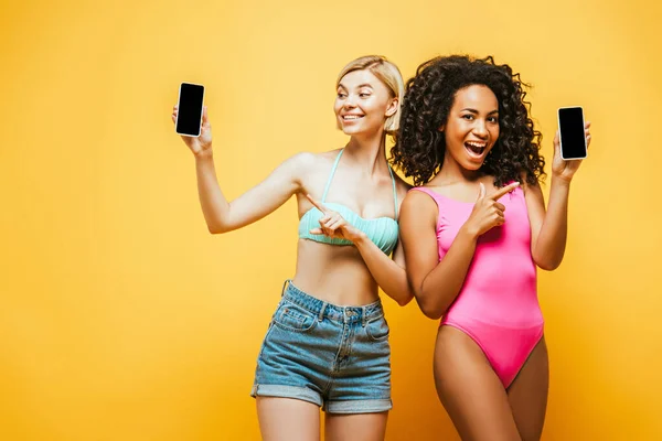 穿着夏装的兴奋的跨种族朋友们指着黄色屏幕上的智能手机 — 图库照片