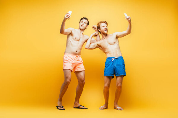 full length view of two shirtless friends demonstrating biceps and grimacing while taking selfie on smartphones on yellow