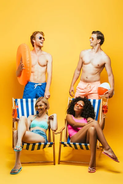 Şişme yüzüklü ve toplu tişörtsüz erkekler çok ırklı kadınların yanında oturmuş, sarı bardaklarda kokteyl kadehleriyle.