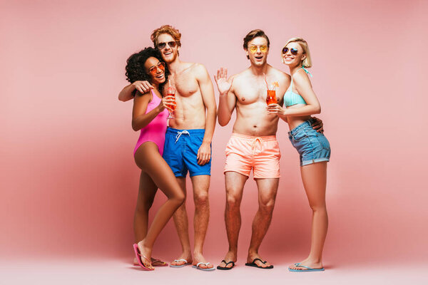 shirtless man waving hand while looking at camera together with multicultural friends holding cocktails isolated on pink