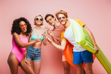 Genç çok kültürlü yaz arkadaşları pembe kameraya bakarken şampanya kadehlerini tokuşturuyorlar.