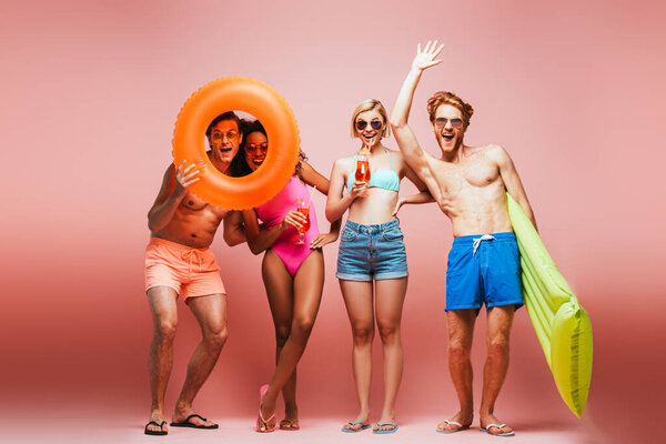 shirtless man waving hand near excited multicultural friends with swim ring and cocktail glasses isolated on pink