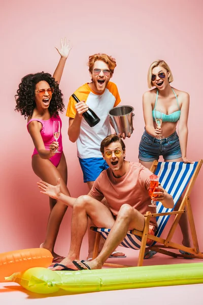 excited man sitting in deck chair with cocktail near multicultural friends with champagne bottle and bucket on pink