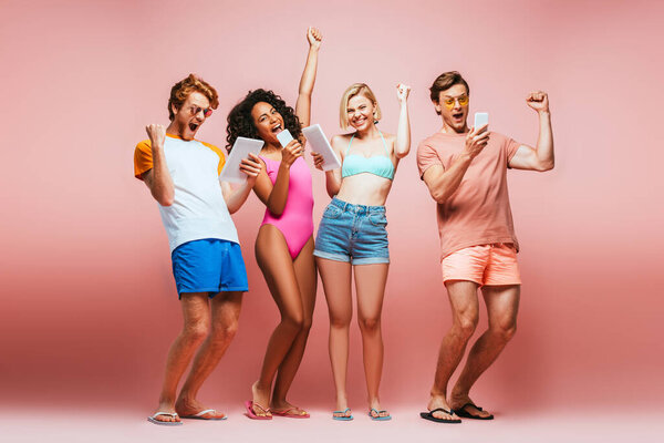 full length view of excited multicultural friends in summer outfit showing winner gesture while looking at gadgets on pink