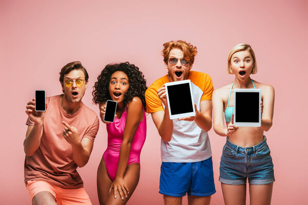 excited multicultural friends in summer outfit showing gadgets with blank screen isolated on pink
