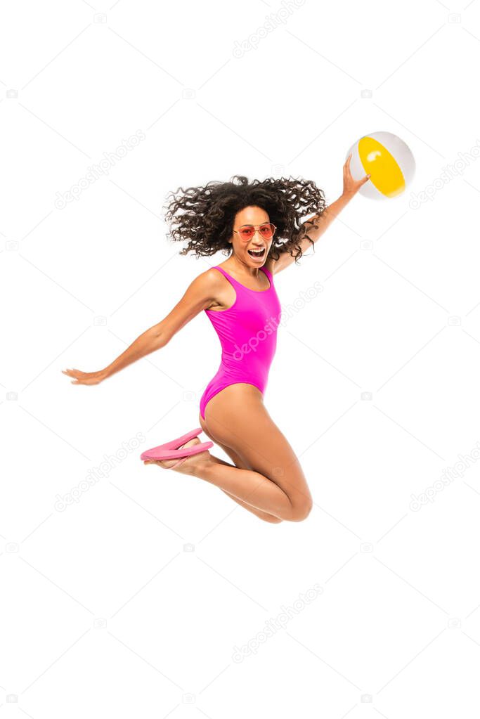 African american woman in sunglasses and swimsuit holding inflatable ball and jumping isolated on white