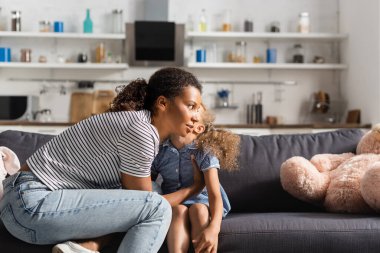 young african american mom in striped t-shirt near daughter whispering in her ear on couch in kitchen clipart