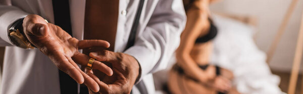 Панорамный урожай мужчины в рубашке снимающего обручальное кольцо с женщиной в нижнем белье на кровати на заднем плане 