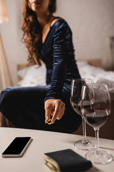 Cropped view of young woman putting wedding ring on coffee table near glasses of wine, smartphone and wallet in bedroom 