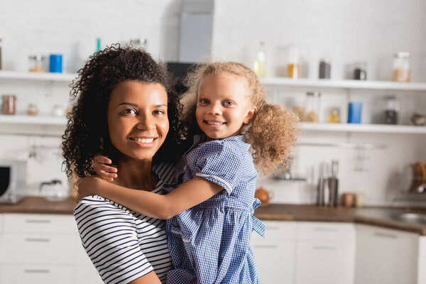 молодая африканская американка держит дочь на руках и смотрит в камеру на кухне