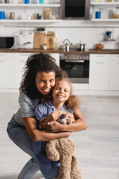 молодая африканская американка обнимает взволнованную дочь с плюшевым мишкой, глядя в камеру на кухне