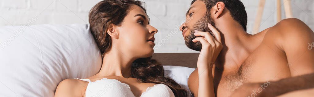 Panoramic shot of woman in bra touching chin of muscular boyfriend on bed 