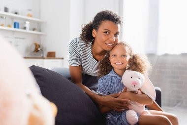 Seçici odak noktası çizgili tişörtlü Afro-Amerikan kadın ve oyuncak tavşanlı kız kameraya bakıyor. 