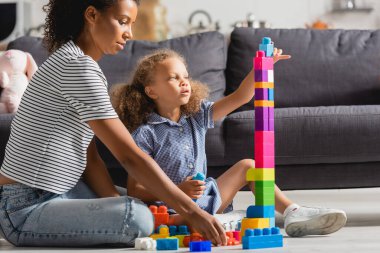 african american child sitting on floor near babysitter and building tower from multicolored blocks clipart