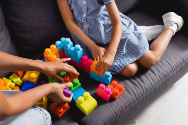 partial view of african american nanny and child playing with multicolored building blocks on sofa clipart