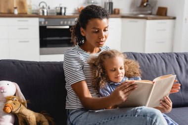 african american girl and young babysitter in striped t-shirt reading book in kitchen  clipart