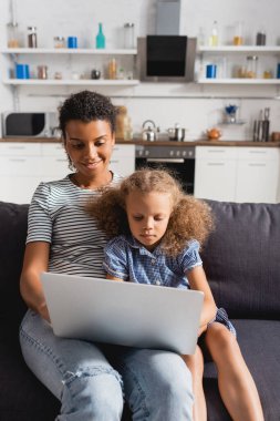 Genç Afrikalı Amerikalı, kızıyla mutfaktaki kanepede otururken dizüstü bilgisayarda çalışıyor.