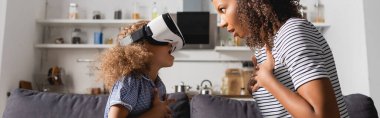 Heyecanlı Afro-Amerikan bebek bakıcısı ve vr kulaklıklı çocuk birbirlerine bakarken göğüslerinin üstünde el ele tutuşuyorlar, panoramik çekim