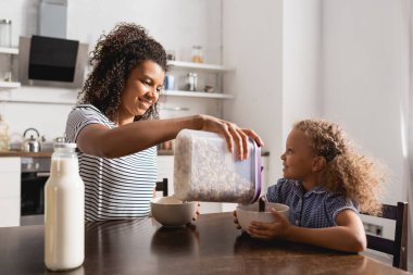 Çizgili tişörtlü Afro-Amerikan annesi kase içine mısır gevreği ve bir şişe taze süt döktü.