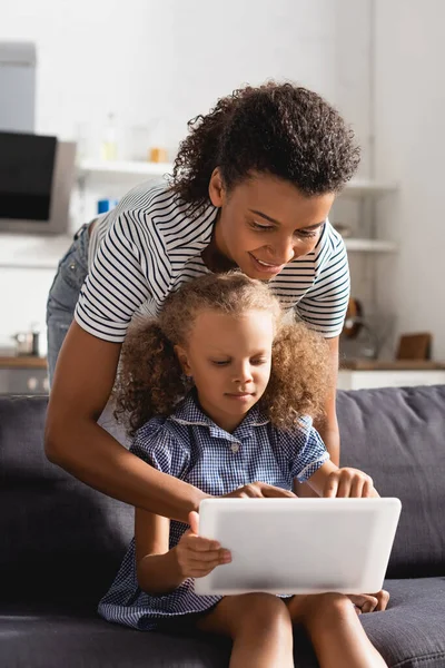 若いアフリカ系アメリカ人のベビーシッターがソファに座っている女の子と一緒にデジタルタブレットを使って — ストック写真