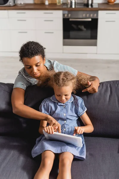 アフリカ系アメリカ人の乳母がデジタルタブレットで指を指しソファに座っている子供の手の中に — ストック写真
