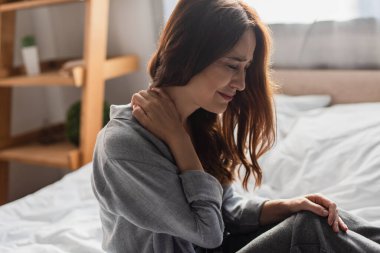 upset brunette woman suffering from neck pain in bedroom clipart