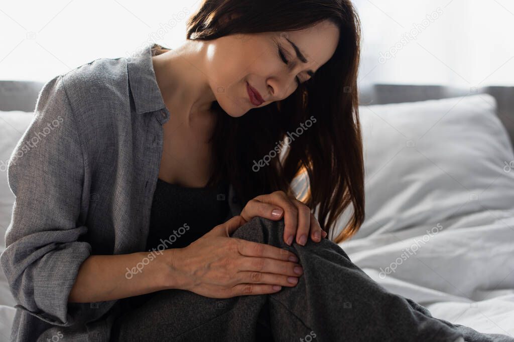 brunette woman suffering from pain in knee while sitting on bed at home