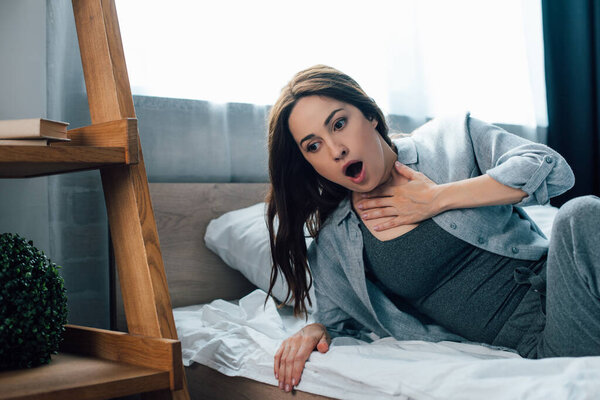 испуганная брюнетка женщина трогает горло в спальне 