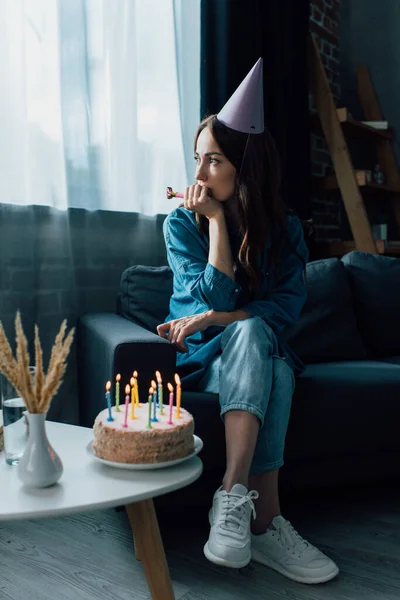 悲しい女がパーティーホーンを持ってコーヒーテーブルの上で誕生日ケーキの近くのソファに座って — ストック写真