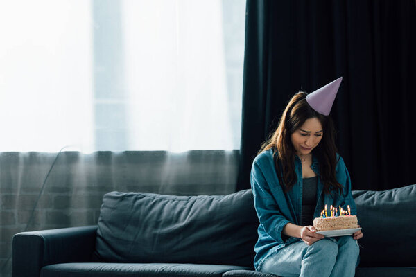 frustrated brunette woman in party cap sitting on sofa and holding birthday cake with candles 