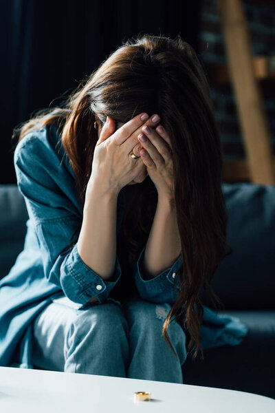 подавленная женщина покрывает лицо сидя рядом с журнальным столиком с золотым кольцом, концепция развода 