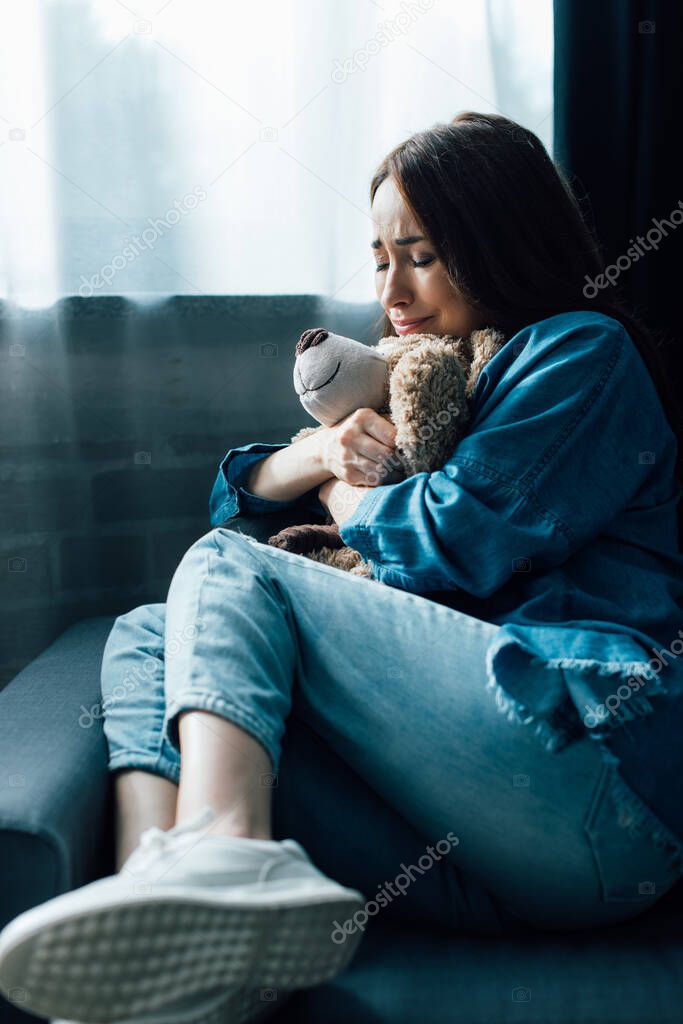 upset brunette woman with closed eyes holding teddy bear while sitting on sofa