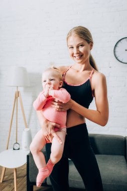 Spor kıyafetli sağlıklı bir kadın oturma odasında kucağında bebek tutuyor. 
